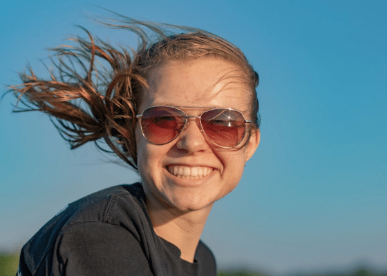 Megan Wolowiec enjoying the summer sun; close-up of smiling woman, wearing sunglasses, long hair blowing in the wind. Photo by Katelyn Wolowiec