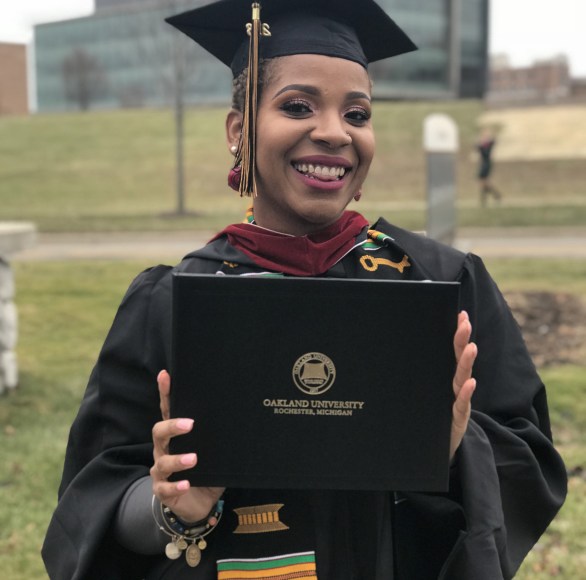 Young African American woman in cap and gown, holding Oakland University diploma, smiles.