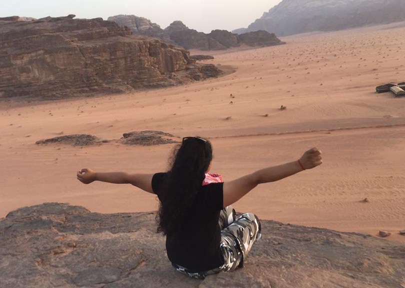 Woman sits on rock above desert line, cliffs in the background; she faces away, arms stretched wide.