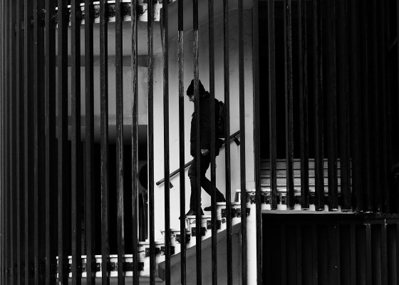Black-and-white image of man with hat and backpack going down stairs, behind decorative bars.