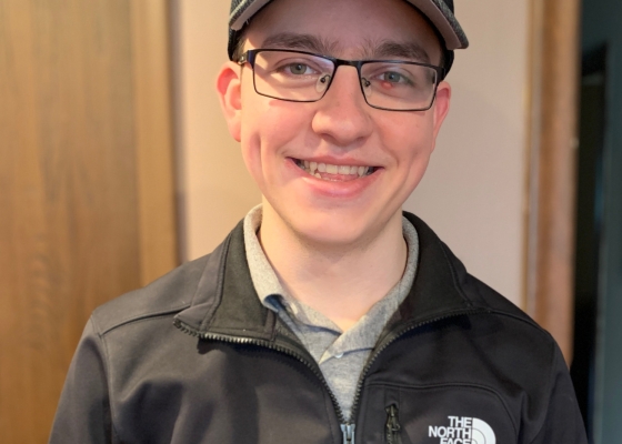 Young adult man smiles to camera, wearing black sweatshirt and baseball cap with Detroit logo on it.