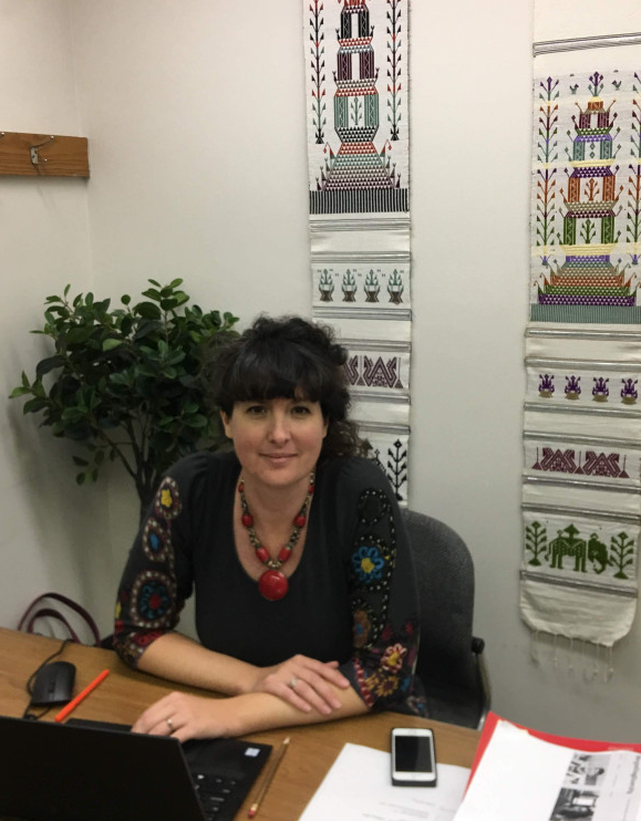 Woman sitting at desk, fabric hangings on the wall behind, laptop, papers, and phone in front of her on the desk.