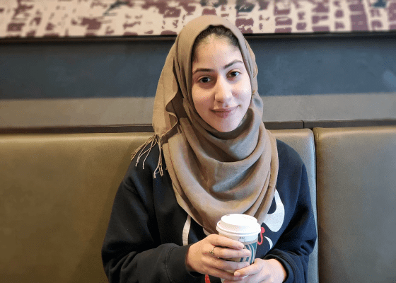 Woman smiles at camera, holds cup of coffee, wears brown head scarf.