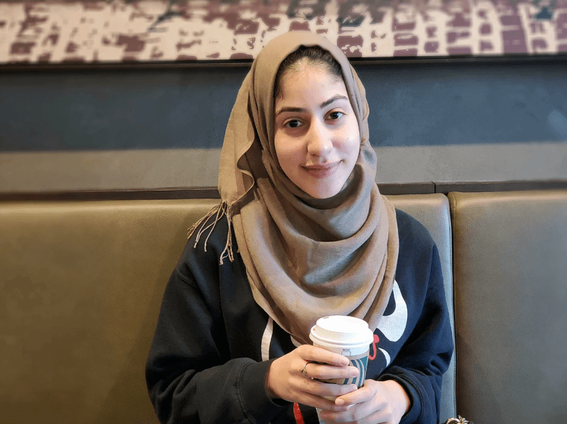 Woman smiles at camera, holds cup of coffee, wears brown head scarf.