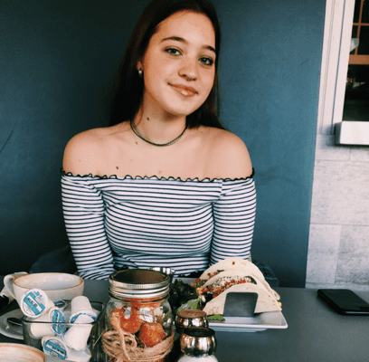 Young woman smiles, sitting at table, food in front of her.