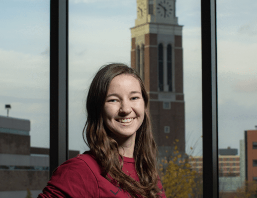 Young woman smiles, Oakland University's clock tower is reflected in the glass window behind her