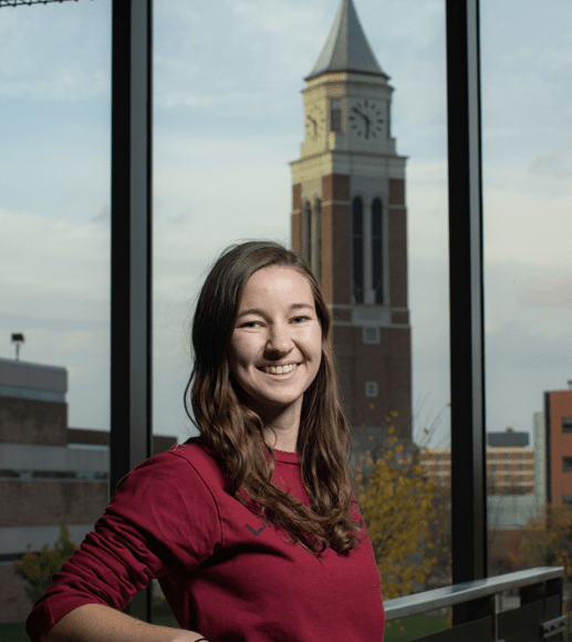 Young woman smiles, Oakland University's clock tower is reflected in the glass window behind her