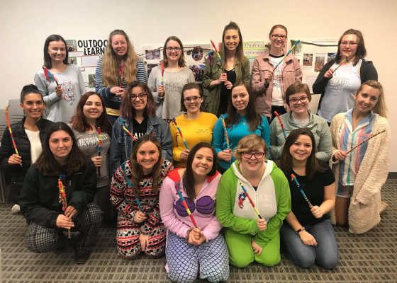 Group of 18 students smile, holding deocrated sticks, some dressed in pajamas.