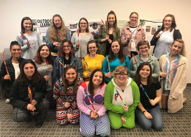 Group of 18 students smile, holding deocrated sticks, some dressed in pajamas.