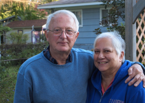 An elderly couple smile, his arm around her shoulders, view of a backyard and blue house behind them.