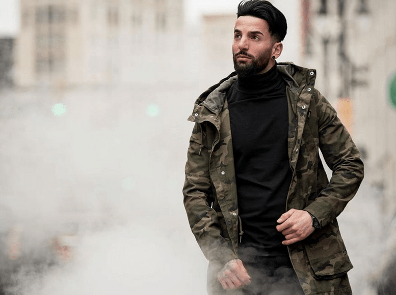 Young man dressed in black and green jacket walks on urban street, smoke coming out of a manhole.