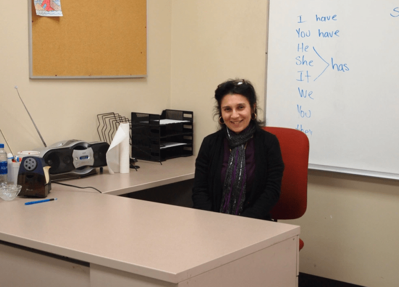 Woman smiles while sitting at desk, whiteboard behind her reading list of pronouns and conjugation of verb "to have."