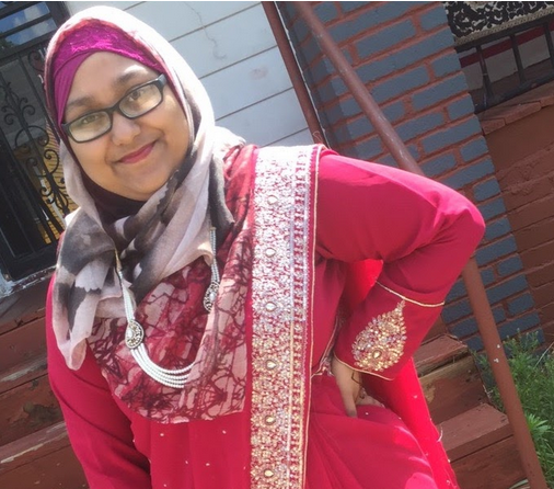 Young woman wearing glasses smiles to camera, as she poses with a red sari and grey and pink head scarf.