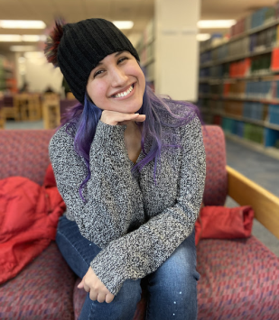 Young woman smiles, sits on couch with book shelves behind, wears hat.