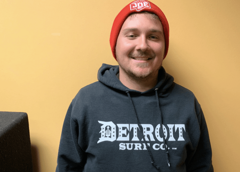 Young man with red hat and "Detroit Surf Co." grey sweatshirt smiles with yellow wall behind him.