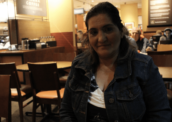 Woman looks at camera, coffee shop setting behind her - counter with coffee canisters, people at tables.
