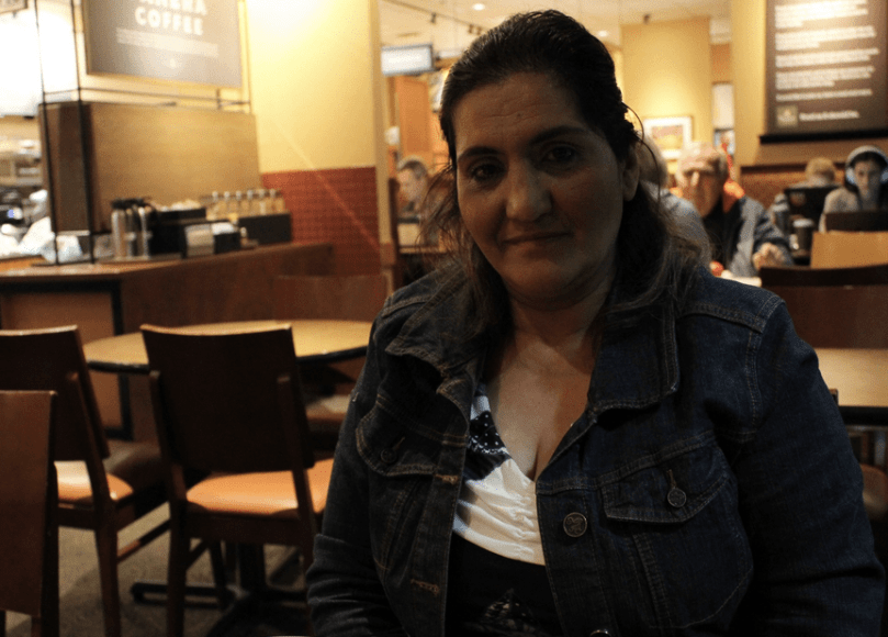 Woman looks at camera, coffee shop setting behind her - counter with coffee canisters, people at tables.