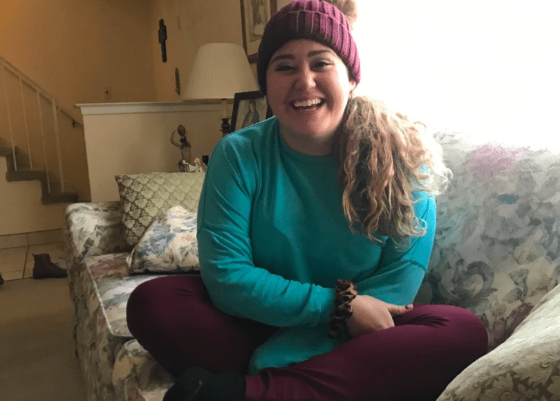 Young woman smiles, sitting on a flowery couch, hat on her head, long hair down on one shoulder.