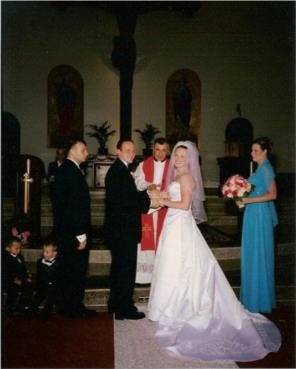Wedding setting, bride and group, framed by maid of honor on one side and groomsmen and two small children on the other side, officiating priest in the background, church setting