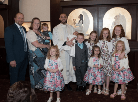 Family poses for picture, parents holding small child on left, six girls in matching outfits, two boys in matching outfits, priest in the center holding baby dressed in white, Catholic church setting