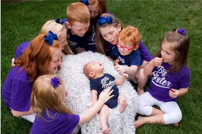 Eight children (six girls and two boys) in matching purple and dark blue shirts look on to small baby in the center. Baby's onesie reads "Little Brother," one of the purple shirts reads "Big Sister."