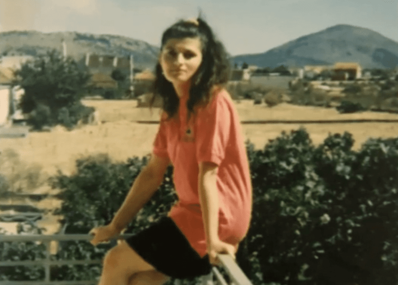 Young woman with dark hair sits on rail with hills and trees in the background