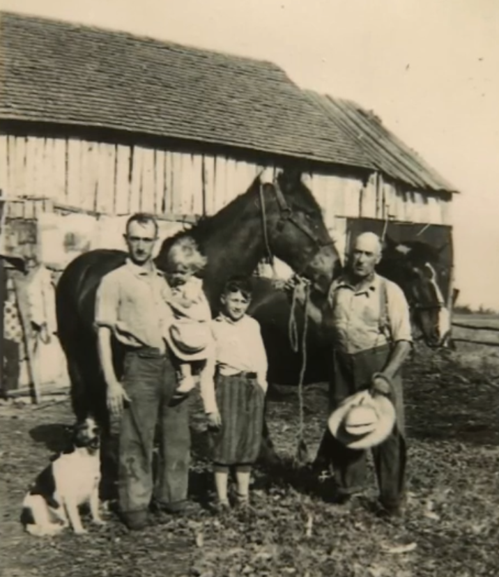 Black-and-white photo of two men, two children, a dog, and two horses in front of a barn.