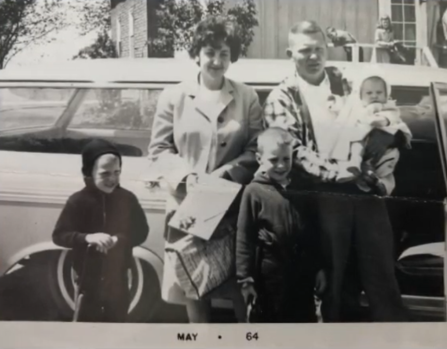 Black-and-white photo of two white parents holding three smaller children, one in the dad's arms, smiling and posing by a car.