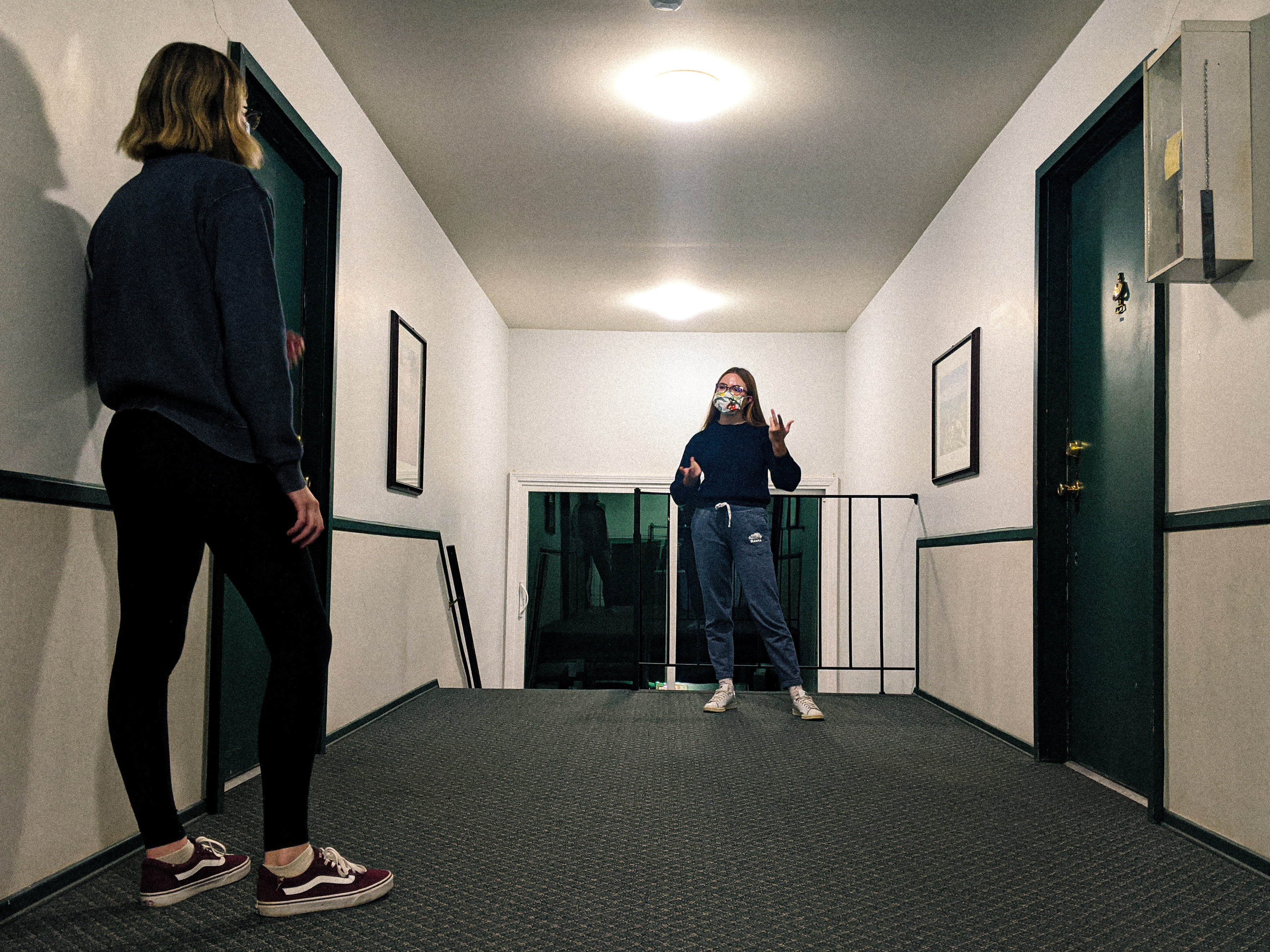 Two young women in a hallway, facing each other from at least six feet distance, wearing face masks.