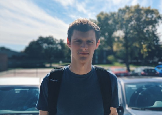 Young white man in blue tshirt, wearing black backpack stands with parking lot behind
