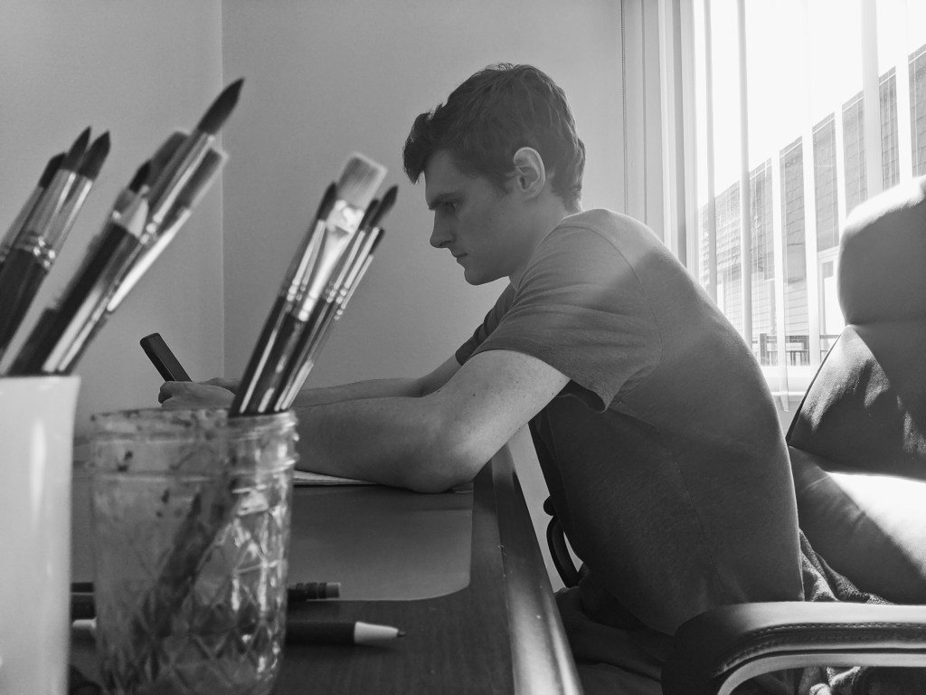 Black and white picture of young man sitting at desk, jars of paint brushes in the foreground