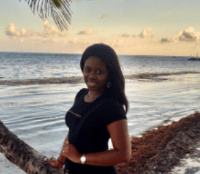 Young woman with dark skin, smiles by palm tree on beach, large body of water behind her