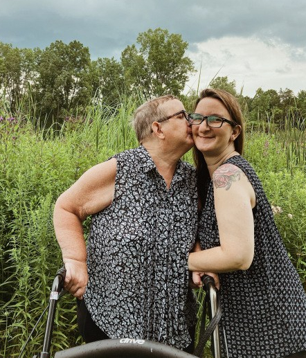 White woman with short grey hair leaning on walker gives a kiss on the cheek to smiling younger woman with long hair