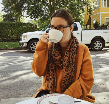 Young white woman with long hair in a ponytail drinks coffee at table outdoors, street view in the background