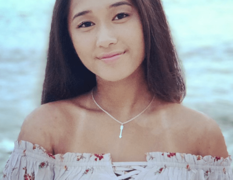 Young Asian woman with long hair smiles in summer dress, body of water behind her