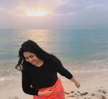 Young white woman with black hair smiles and leans over by large body of water and beach