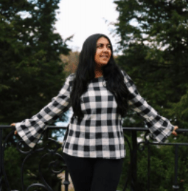Young woman with long, black hair and white-and-black checkered shirt smiles on bridge with trees in the background