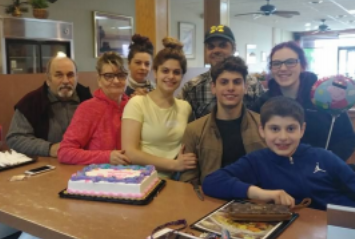 Family smiling at a table, all white with dark hair, of different ages, sheet cake in front of them, one holds a balloon