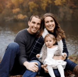 Family of three white people, young daughter on the mom's lap, smile with river in the background