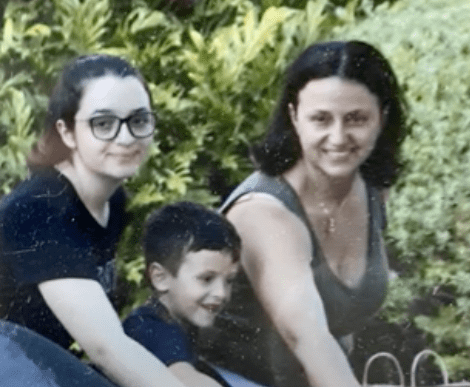 Smiling white woman with black hair next to a teen girl and young boy, with greenery behind them
