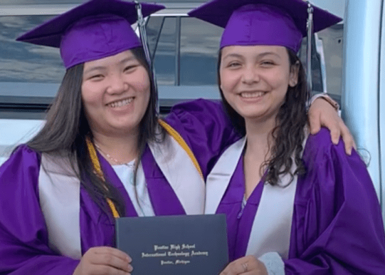 Two young women in graduation robes smile and hold each other, one Asian and the other Latina