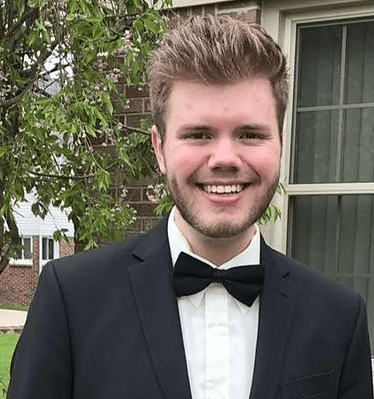 Young, blond, white man with facial hair smiles at camera, wears a tuxedo and bowtie