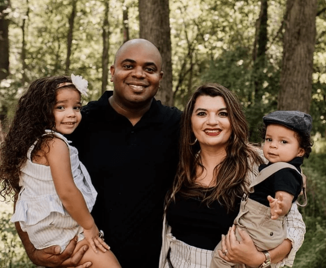 Family of four, black father holds mixed race young daughter, Mexican woman holds young toddler boy, all smile with forest in the background