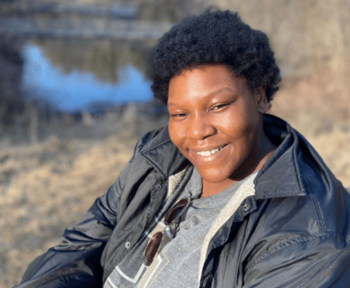 Young Black woman smiles in rain jacket with blurry body of water in the background